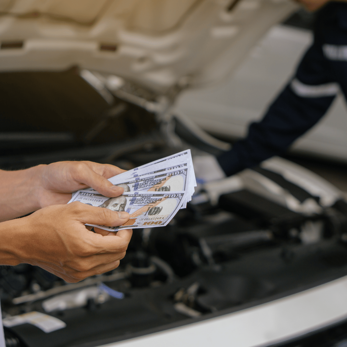 Image of a hand holding several dollar bills, with a mechanic checking a car with hood up in the background.