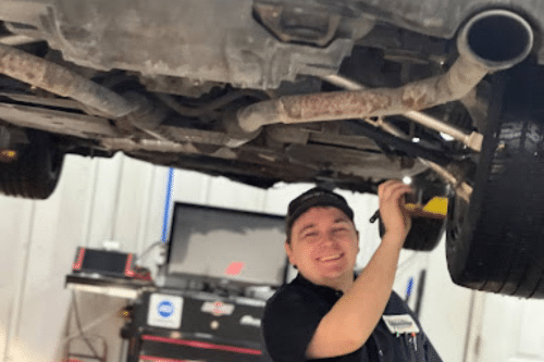 Comprehensive Pre-Trip Inspections | Quality Car Care North Liberty, Iowa. Image of mechanic at Quality Car Care smiling holding flashlight under car on lift in shop doing a pre trip inspection.