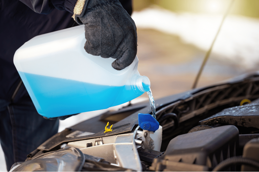 car fluids, auto repair in North Liberty, IA by Quality Car Care. Person wearing gloves pouring blue windshield washer fluid into a car’s reservoir during cold weather.