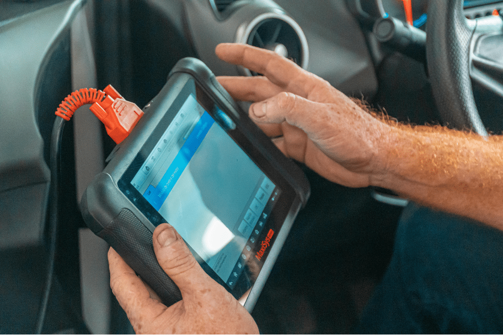 summer car maintenance tips, auto repair in North Liberty, IA by Quality Car Care. Technician uses an advanced handheld diagnostic scan tool connected to a vehicle's OBD port to troubleshoot engine performance issues.